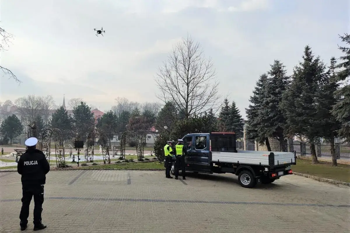 Dron policyjny w powietrzu, obok radiowozu i policjantów.