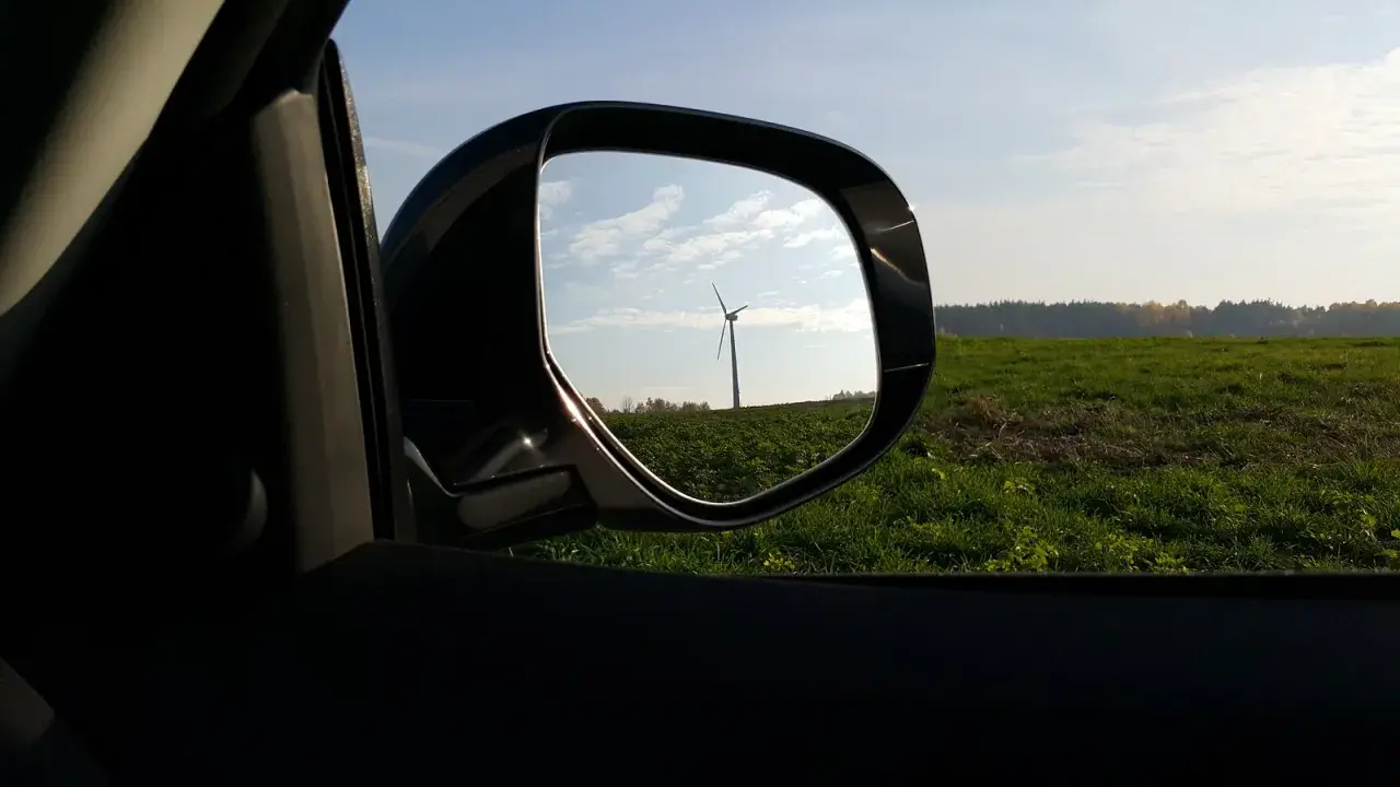 Widok w lusterku samochodowym na wiatrak na tle zielonego pola i nieba.