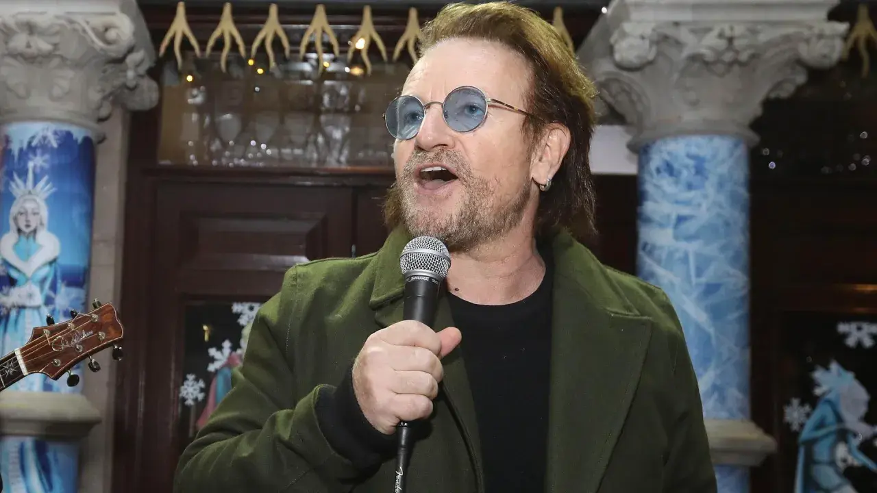 Bono singt ins Mikrofon, trägt eine grüne Jacke und blaue Sonnenbrillen. Der Titel von Bono ist ein bekannter Musiker.