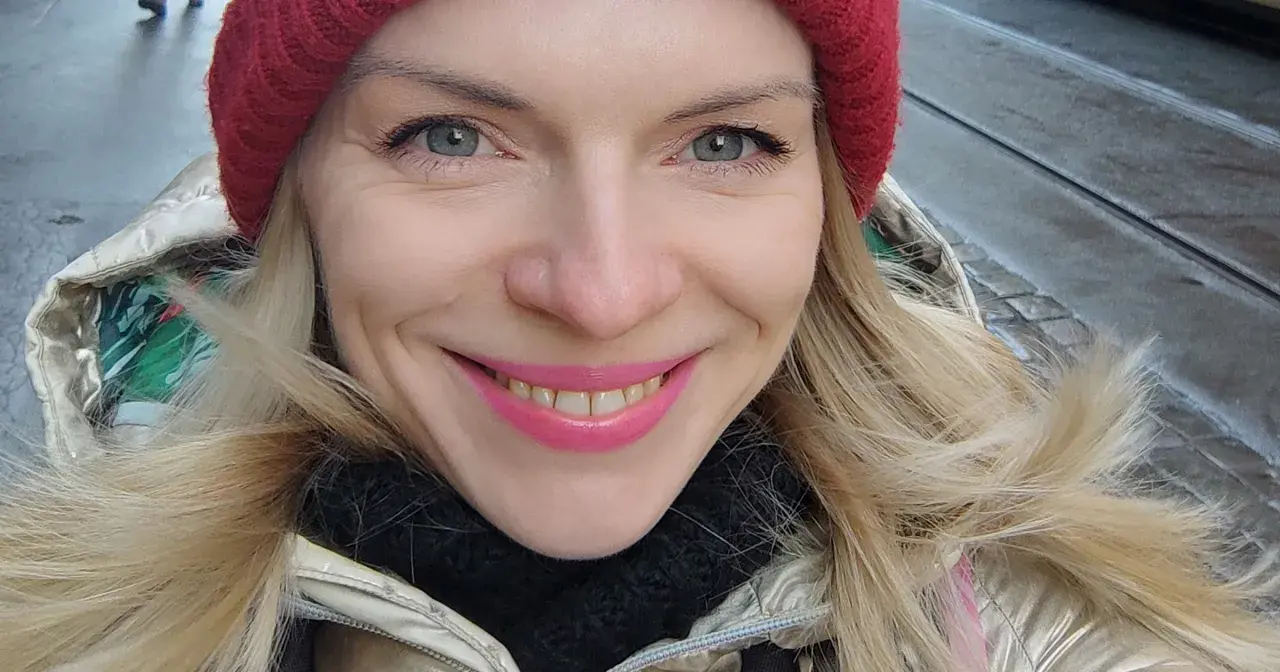 Uśmiechnięta blondynka w czerwonej czapce i złotej kurtce, jakby właśnie spacerowała po ulicach Czech, gdzie ludność cieszy się zimową aurą.
