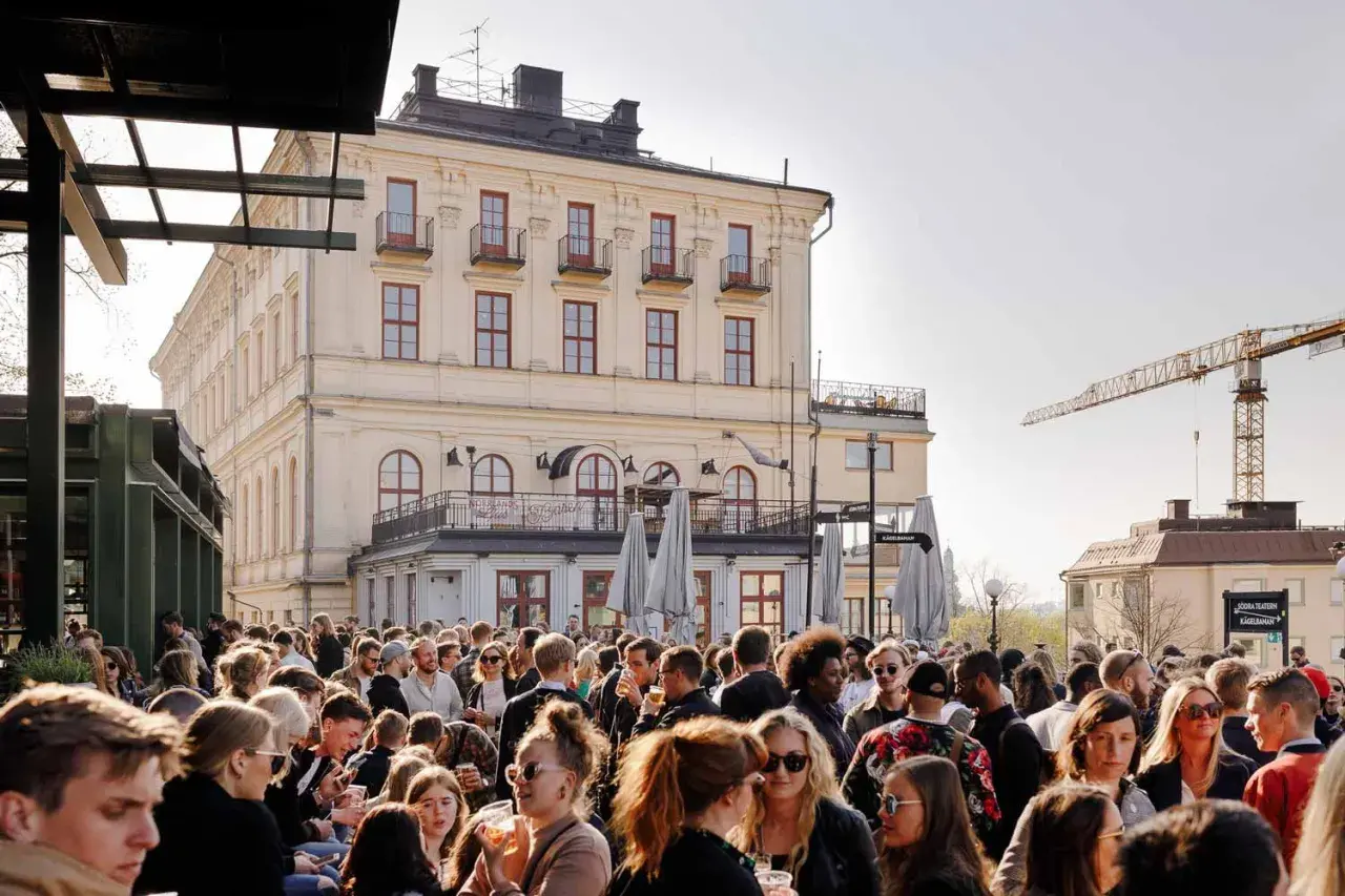 Folk trängs på Mosebacke terassen en solig dag. En stor folkmassa njuter av stämningen framför en gul byggnad.