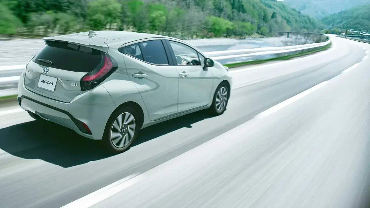 Jasnoszary Toyota Aqua jedzie po krętej drodze wśród zielonych wzgórz.