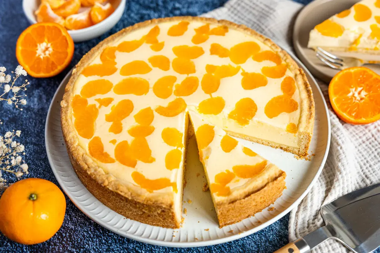 Ein saftiger faule weiber kuchen mit Mandarinenstücken, der auf einem Teller liegt. Daneben liegen frische Mandarinen und eine Tortenschaufel.