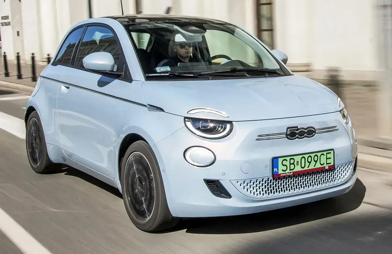 Jasnoniebieski fiat elektryczny 500 z czarnym dachem jedzie ulicą. Kierowca w okularach przeciwsłonecznych prowadzi auto.