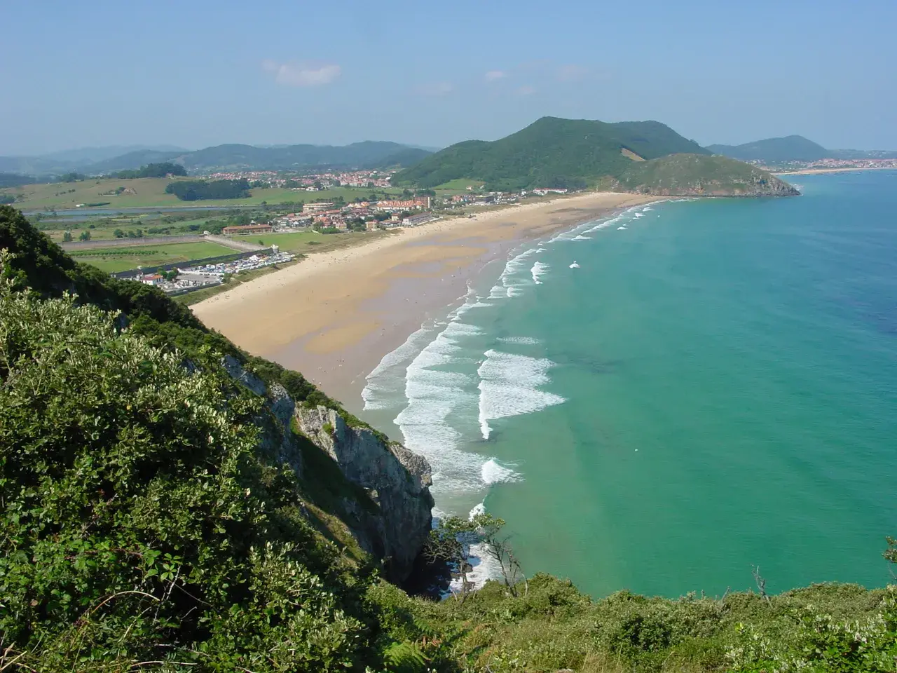 Vista panorámica de la playa de Berria, con arena dorada, aguas turquesas y colinas verdes al fondo.