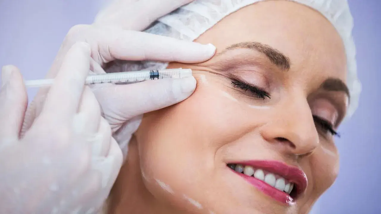 Kobieta uśmiecha się podczas zabiegu baby botox. Dłoń w rękawiczce trzyma strzykawkę z igłą, aplikując preparat w okolicę oka.