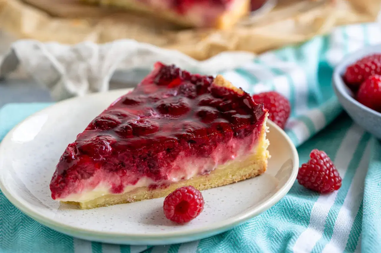 Ein saftiges Stück Himbeerkuchen mit gefrorenen Himbeeren, glasiert und auf einem Teller mit frischen Beeren daneben.