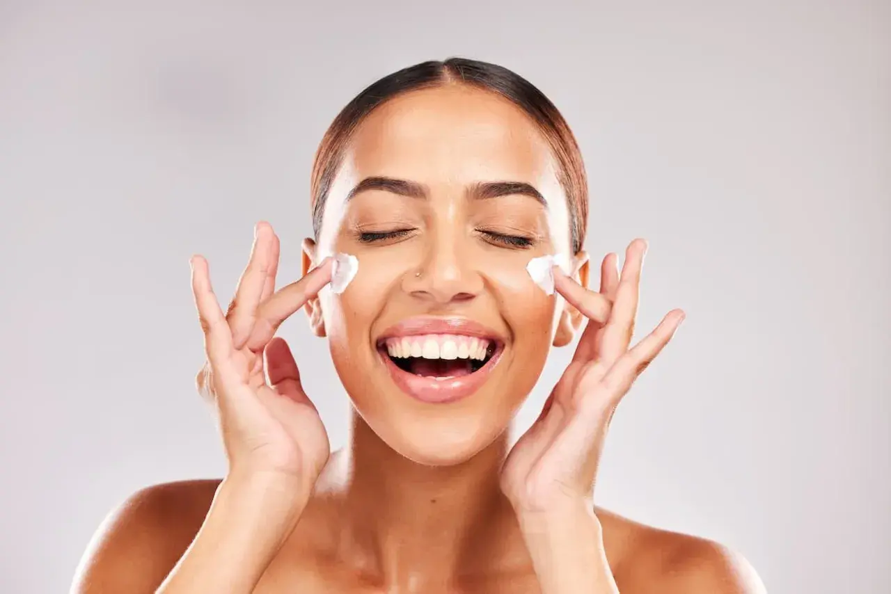 Mujer sonriente aplicándose crema en el rostro. Descubre para qué sirve la niacinamida en la piel y luce radiante.