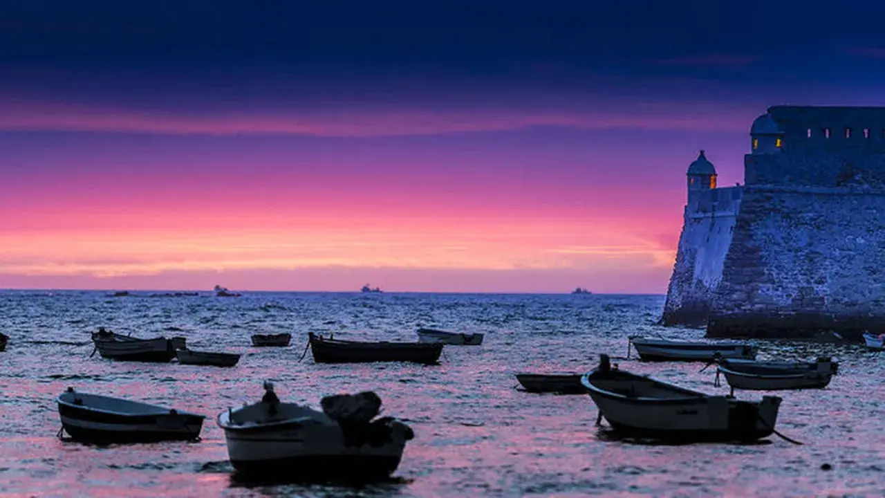 Barcas flotando en el mar al atardecer, con un castillo histórico y un cielo púrpura y rosa. Una hermosa puesta de sol en Cádiz.