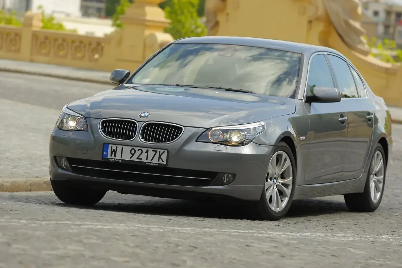 Szary sedan BMW E60 z włączonymi światłami. Widać charakterystyczne żarówki BMW E60, które oświetlają drogę.