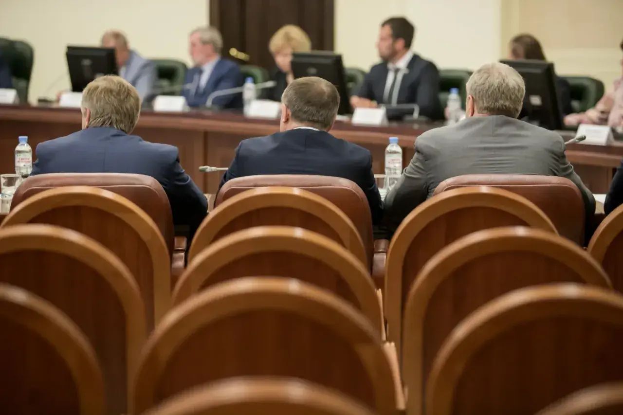 Mężczyźni w garniturach siedzą przy stole, czekając na **absolutorium**. W tle widać monitory i innych uczestników spotkania.
