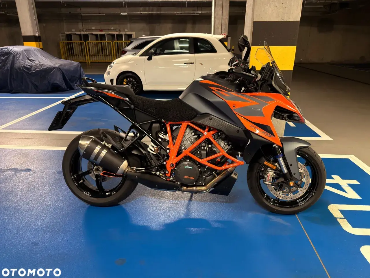 Pomarańczowo-szary KTM 1290 Super Duke w garażu, obok białego Fiata 500.