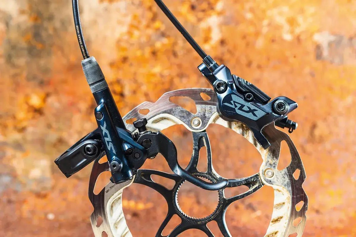 Shimano SLX M7120 hydraulic disc brakes, ready for any trail. The dark blue calipers and rotors are mounted on a rusty, textured background.