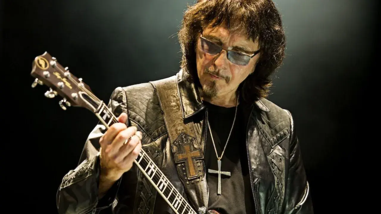 Tony Iommi fingers grają na gitarze, w okularach i skórzanej kurtce z krzyżem.