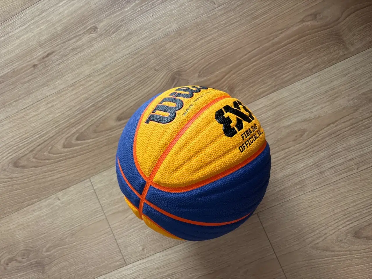 Piłka do koszykówki 3x3 Wilson z pomarańczowo-niebieskimi panelami leży na drewnianej podłodze.