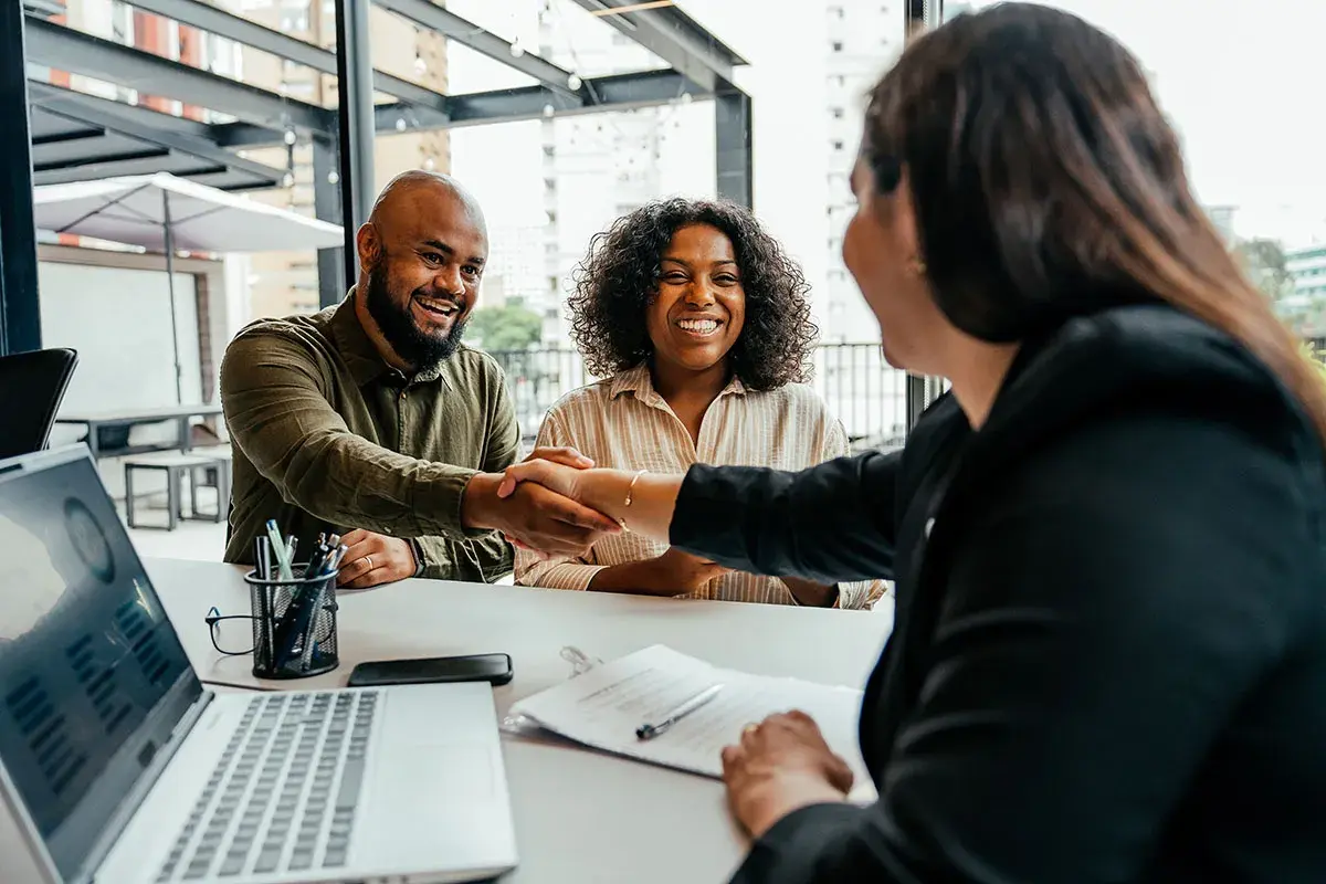 A man and woman smile, shaking hands after successful negotiation classes online. A laptop shows charts.