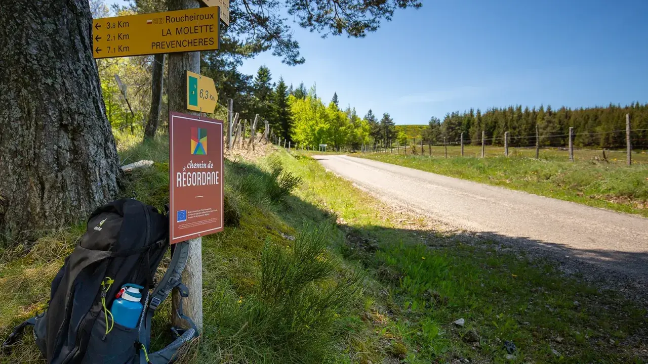 Sac à dos prêt pour l'aventure sur le chemin de Regordane. Panneaux indicateurs et route s'étendent sous un ciel bleu.