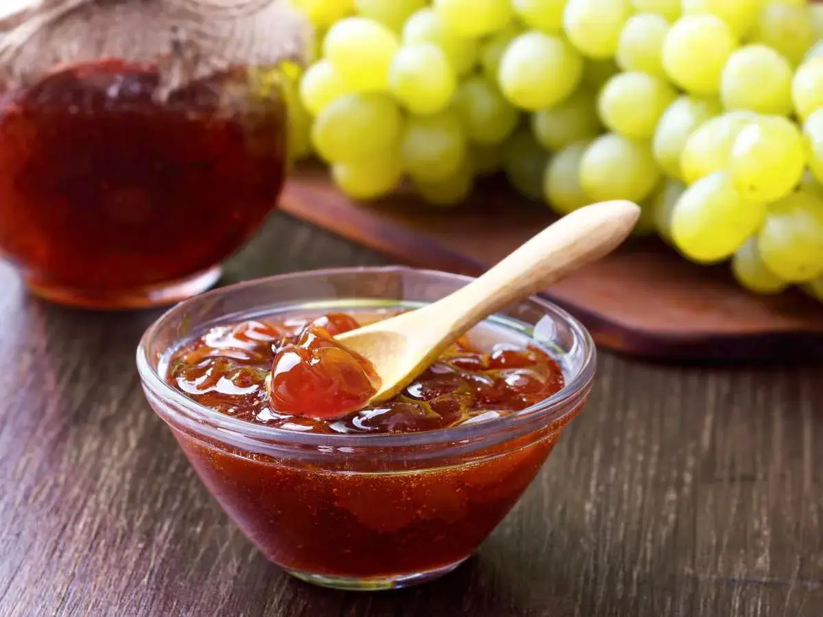 Un cuenco de cristal con mermelada de uvas casera, una cuchara de madera y un racimo de uvas verdes al fondo.