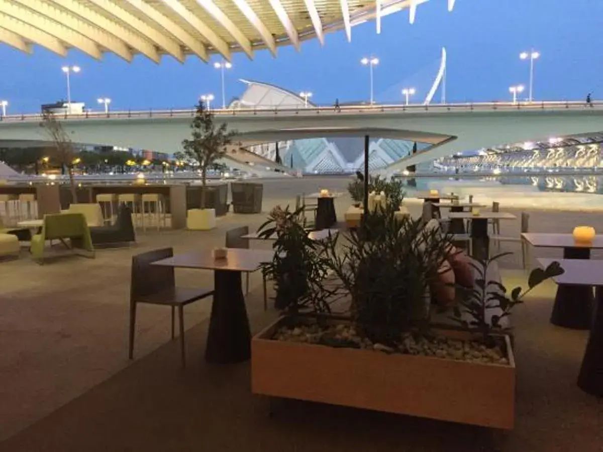 Mesas y sillas en la terraza de restaurantes con vistas a un puente moderno y un cielo azul crepuscular.
