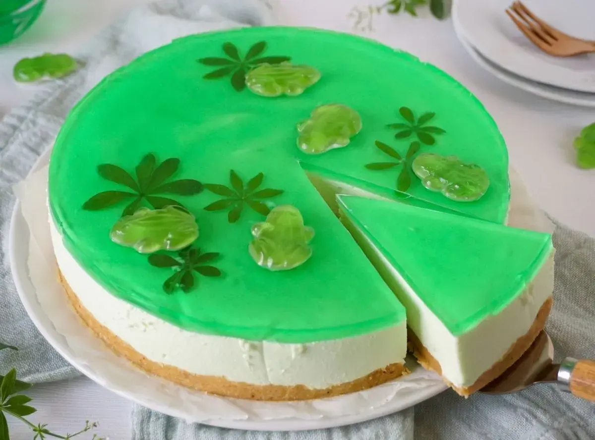 Grüne Philadelphia Torte Waldmeister mit Gummifröschen und Blättern verziert. Ein Stück wird gerade herausgeschnitten.