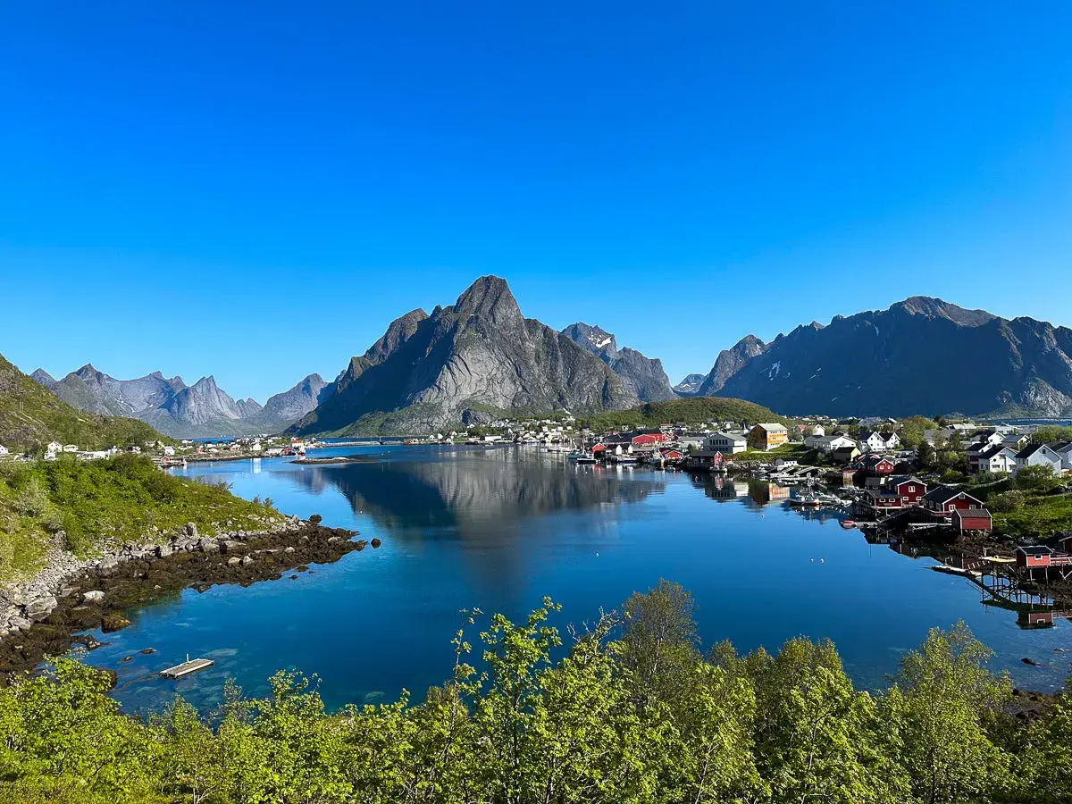 En klarblå himmel speglar sig i det lugna vattnet vid en idyllisk by. Majestätiska berg reser sig bakom de röda husen. En vacker lofoten sommarvy.