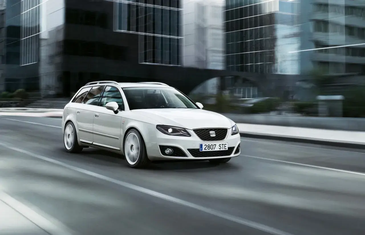 Biały Seat Exeo 2.0 TDI kombi pędzi ulicą miasta, mijając nowoczesne budynki.
