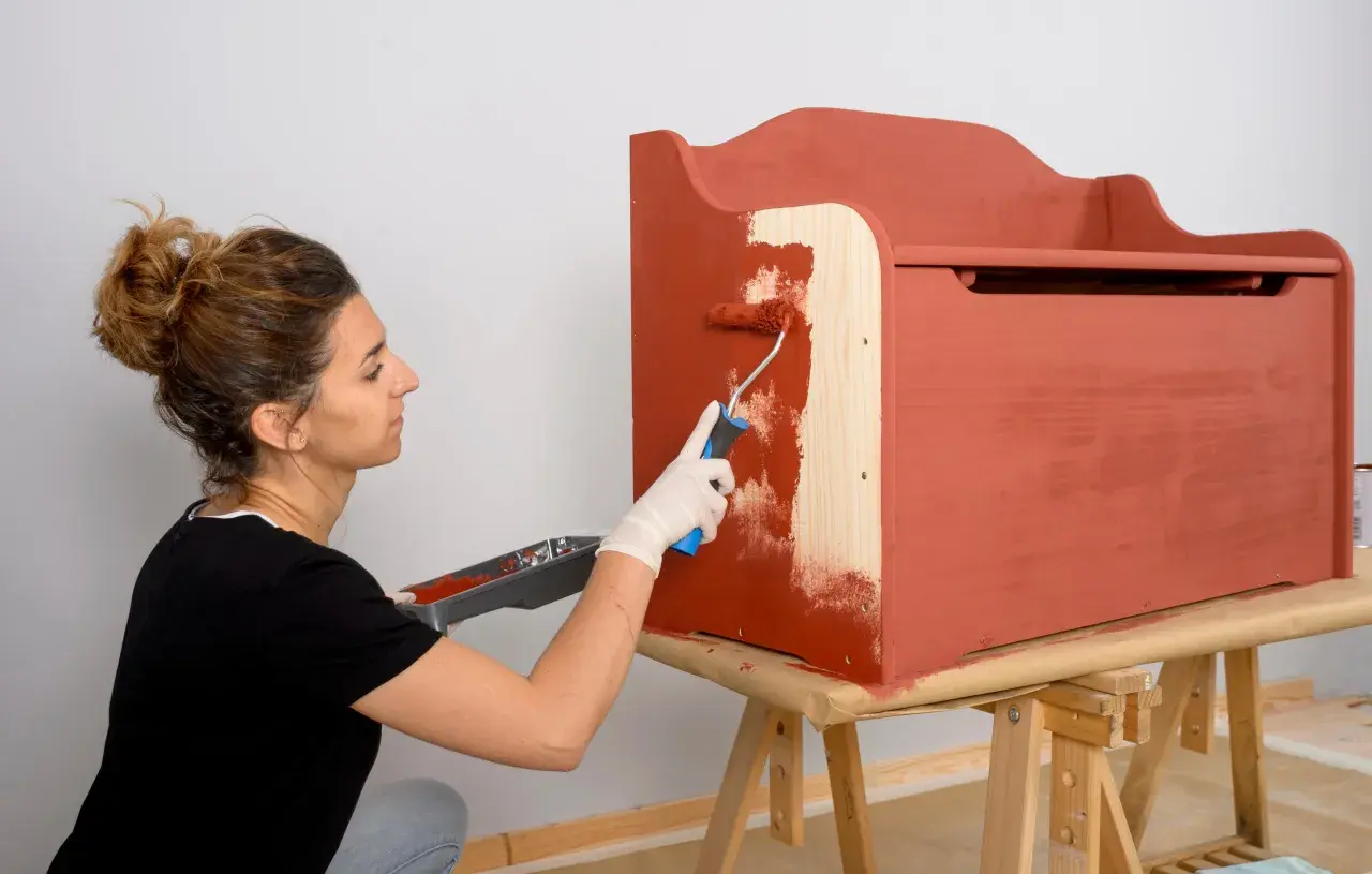 Mujer con guantes blancos pinta con rodillo una caja de madera de color terracota.