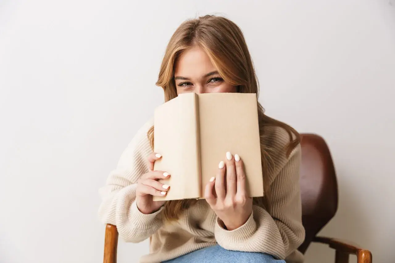 Dziewczyna z uśmiechem ukrywa twarz za książką, zanurzona w świecie literatury young adult.