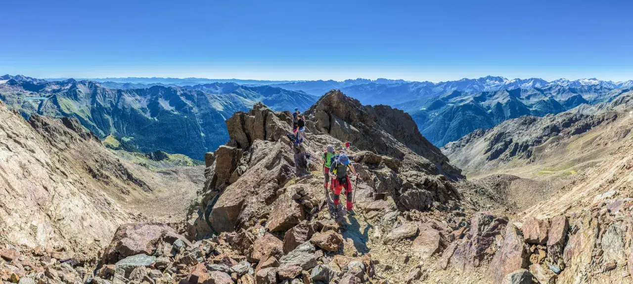Grupa ludzi wyrusza na trekking po skalistym szczycie, podziwiając rozległe pasmo górskie w oddali.