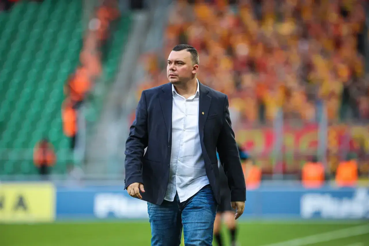 Trener Jagiellonii w eleganckim garniturze i dżinsach, na tle trybun stadionu wypełnionych kibicami.