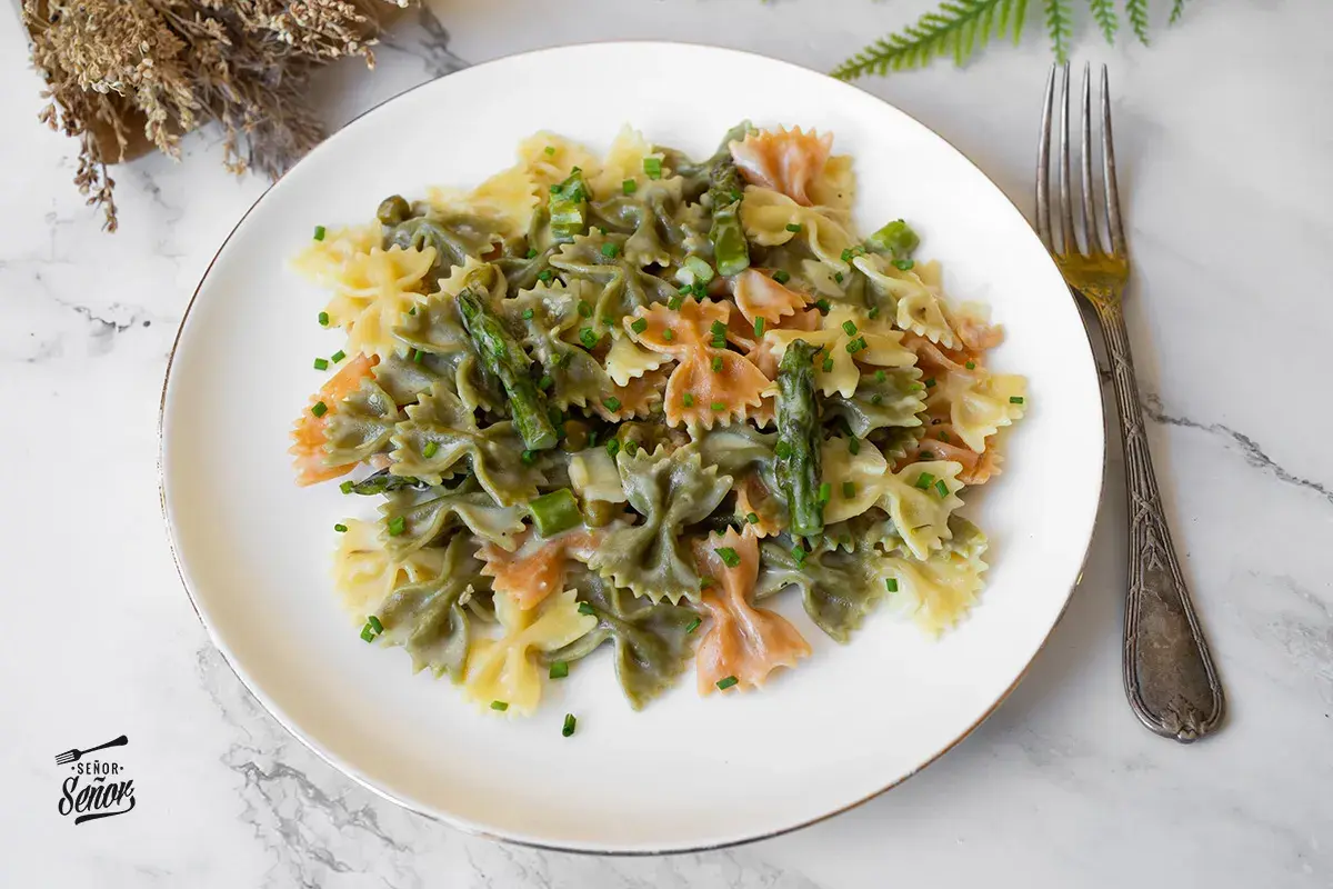 Plato de pasta primavera con farfalle tricolor, espárragos y guisantes en salsa cremosa, adornado con cebollino picado.