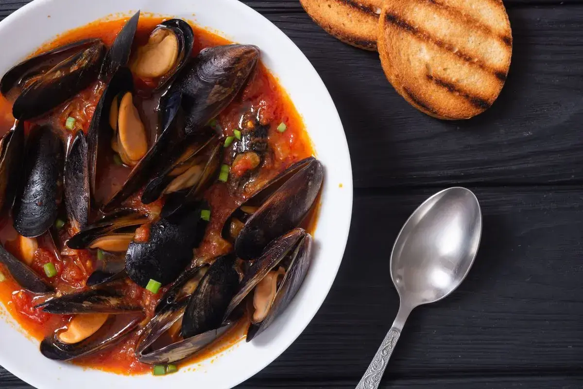 Un invitante piatto di zuppa di cozze in un ricco sugo di pomodoro, guarnito con erba cipollina, accompagnato da crostini tostati e un cucchiaio.