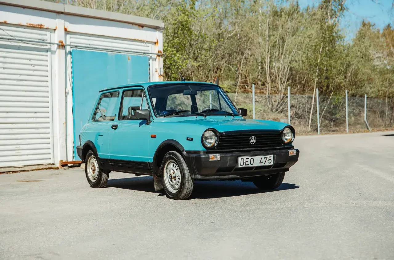 En ljusblå Lancia A112 Junior står parkerad framför en blå port och en vit containerbyggnad.