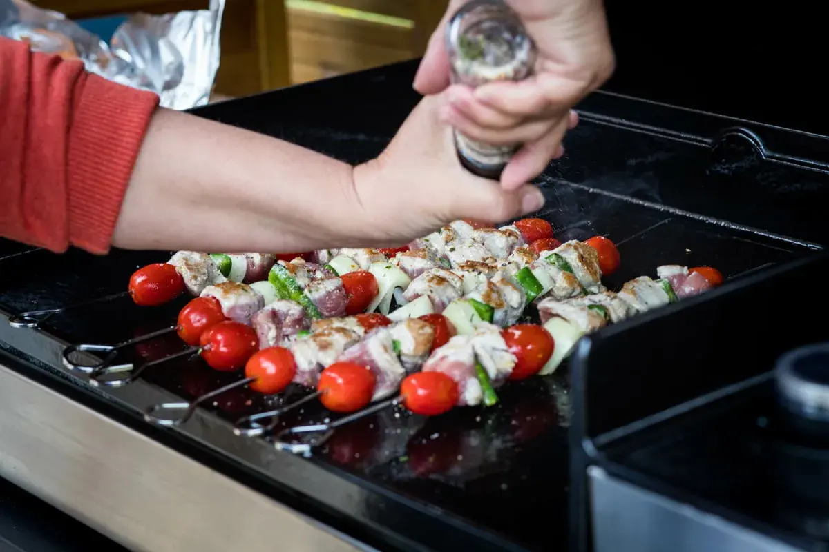 Brochettes de viande, tomates et courgettes sur une plancha à gaz. Une main assaisonne les brochettes.
