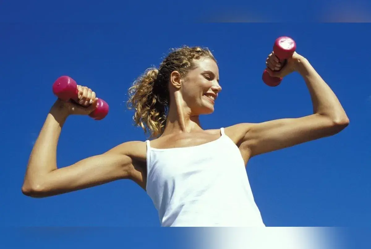 Femme souriante avec des haltères roses, montrant ses muscle bras. Elle porte un débardeur blanc sous un ciel bleu clair.