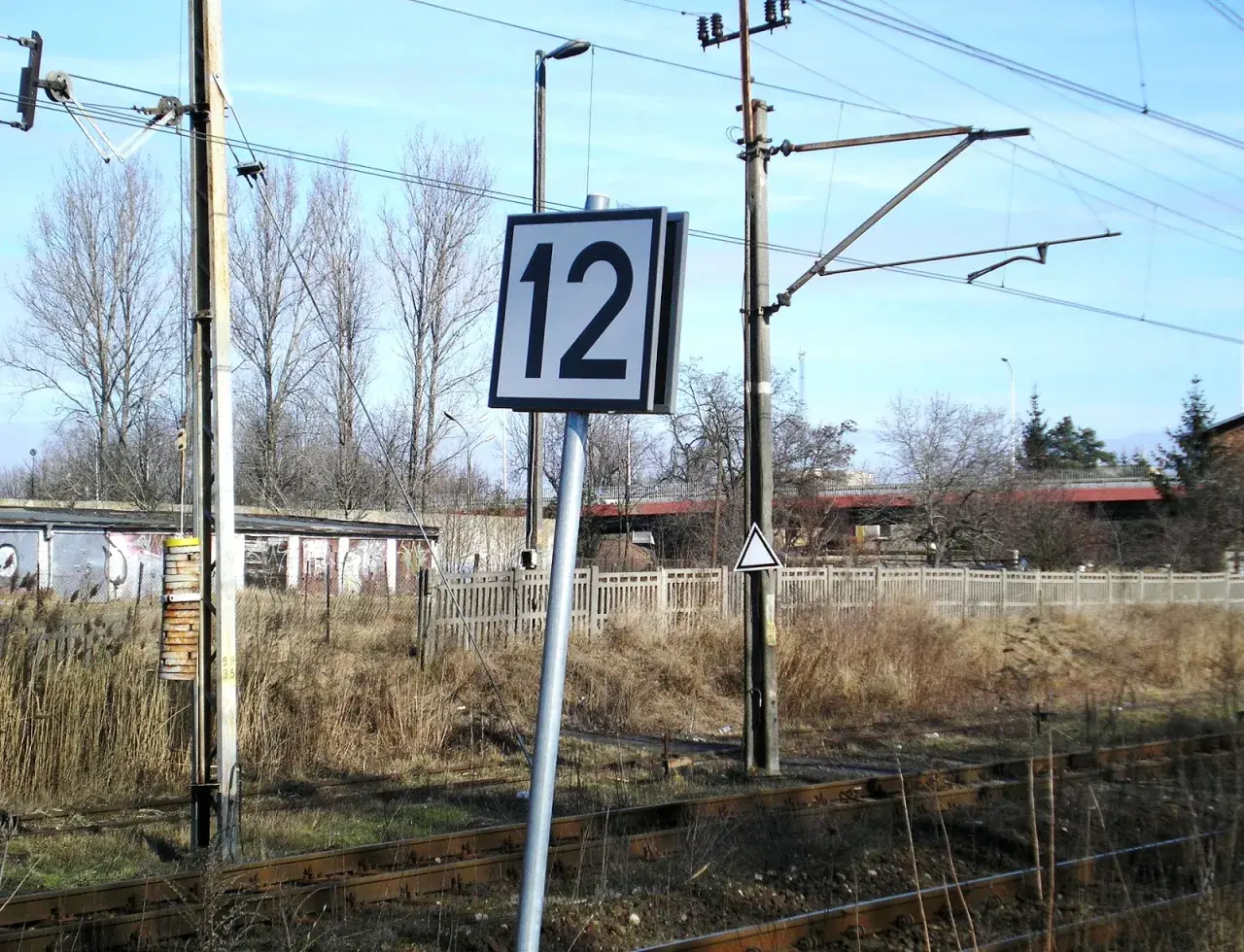 Wskaźnik W24 z numerem 12 przy torach kolejowych. W tle budynki i drzewa.
