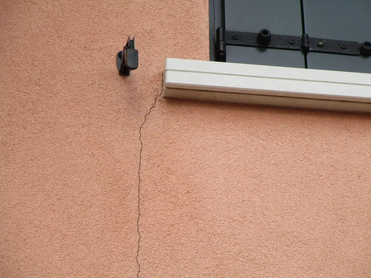 Una crepa sottile attraversa l'intonaco rosa di un muro esterno, vicino a un supporto metallico e a una finestra con persiana. Utile per capire come riempire le crepe nei muri esterni.