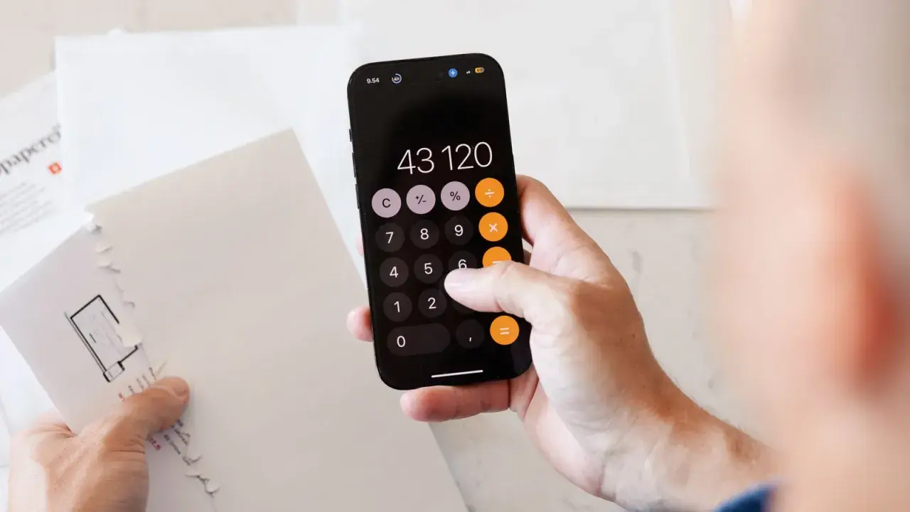 Dłoń z telefonem wyświetlającym kalkulator, na którym widnieje kwota 43120. Obok leżą białe koperty, sugerujące problemy z zadłużeniem.