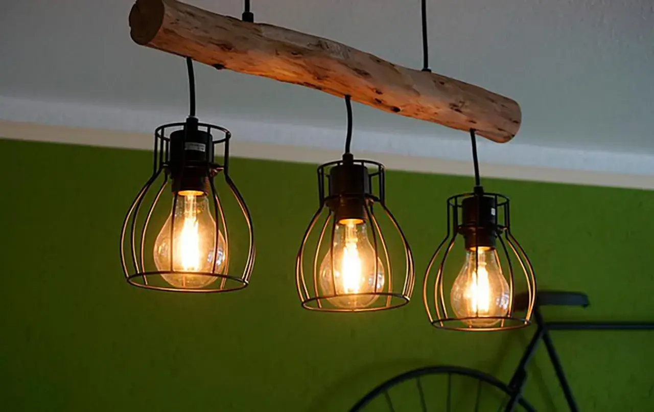 Lámpara de techo colgante con tres bombillas Edison dentro de jaulas metálicas, suspendidas de una viga de madera rústica.