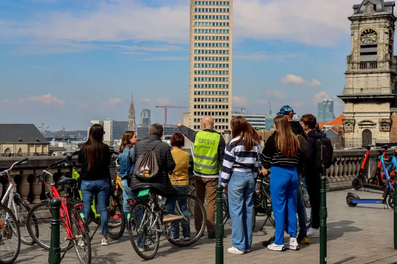 A group gathers for Brussels bike tours, with a guide pointing towards the city skyline featuring a tall skyscraper and an old clock tower.