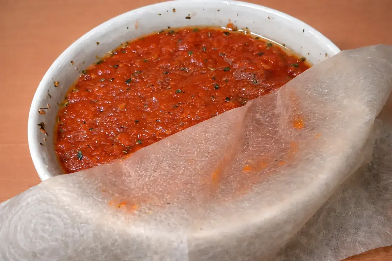 Un tazón blanco con la mejor salsa de tomate para pizza, con hierbas y un trozo de papel de arroz.