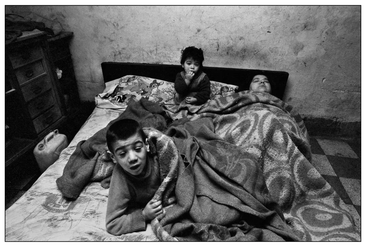 Bambini in un letto, un piccolo è seduto con le dita in bocca, un altro è sdraiato. Un'immagine toccante, una delle **Letizia Battaglia foto famose**.