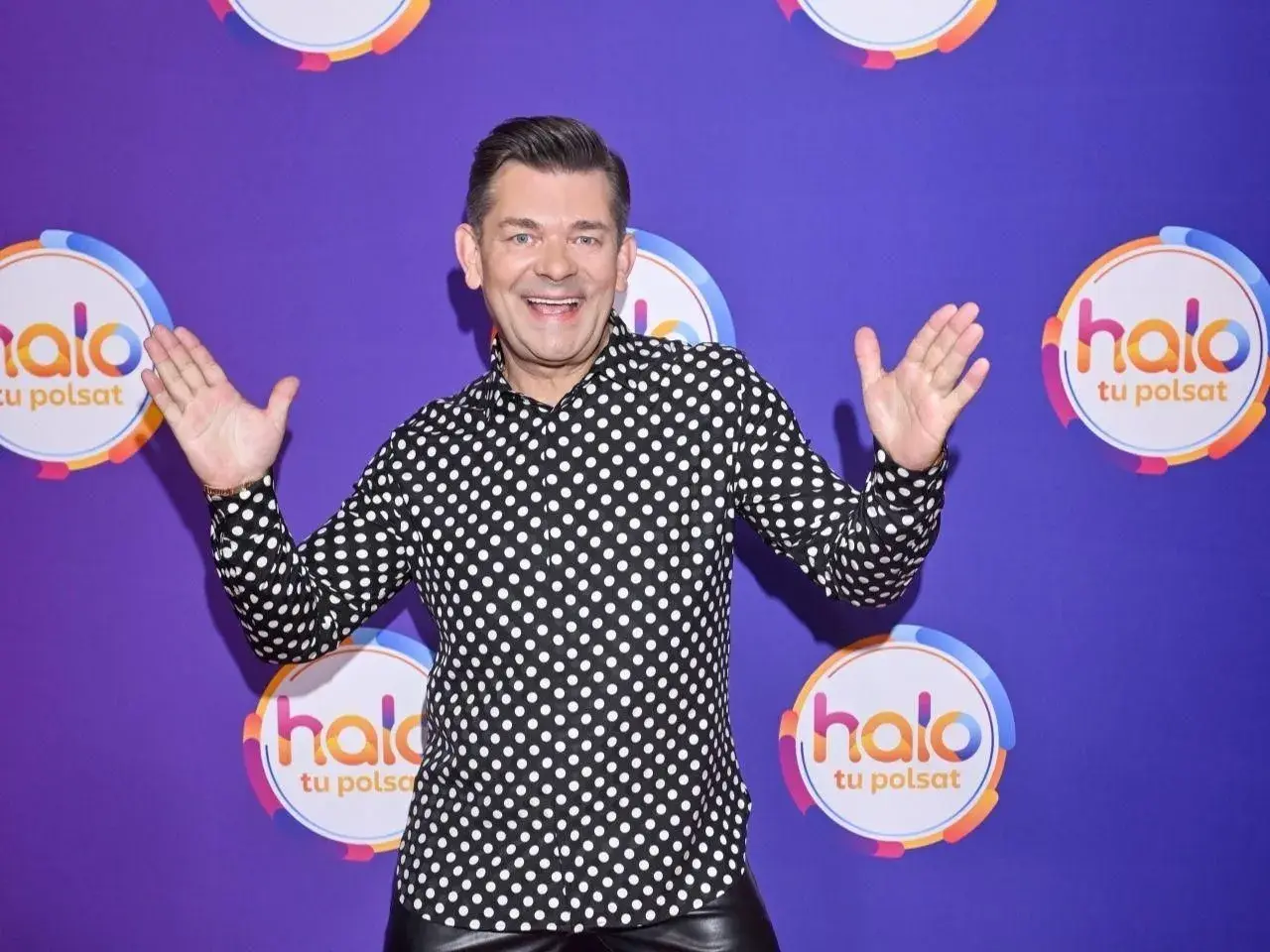 Zenek Martyniuk w czarno-białej koszuli w grochy, z podniesionymi rękami, na tle fioletowym z logo "halo tu Polsat".