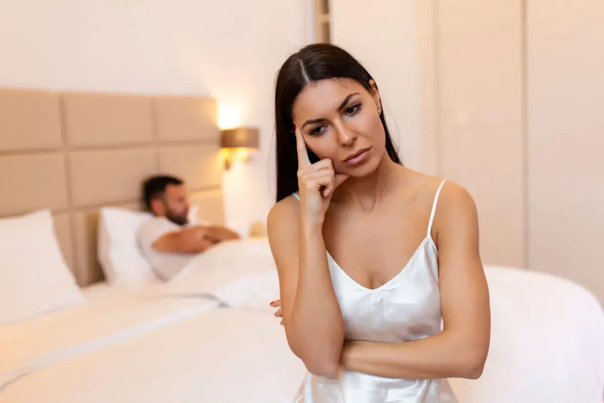 Donna pensierosa in camera da letto, mentre l'uomo è a letto. Potrebbe essere un momento per capire come aumentare la voglia di fare l'amore nell'uomo.
