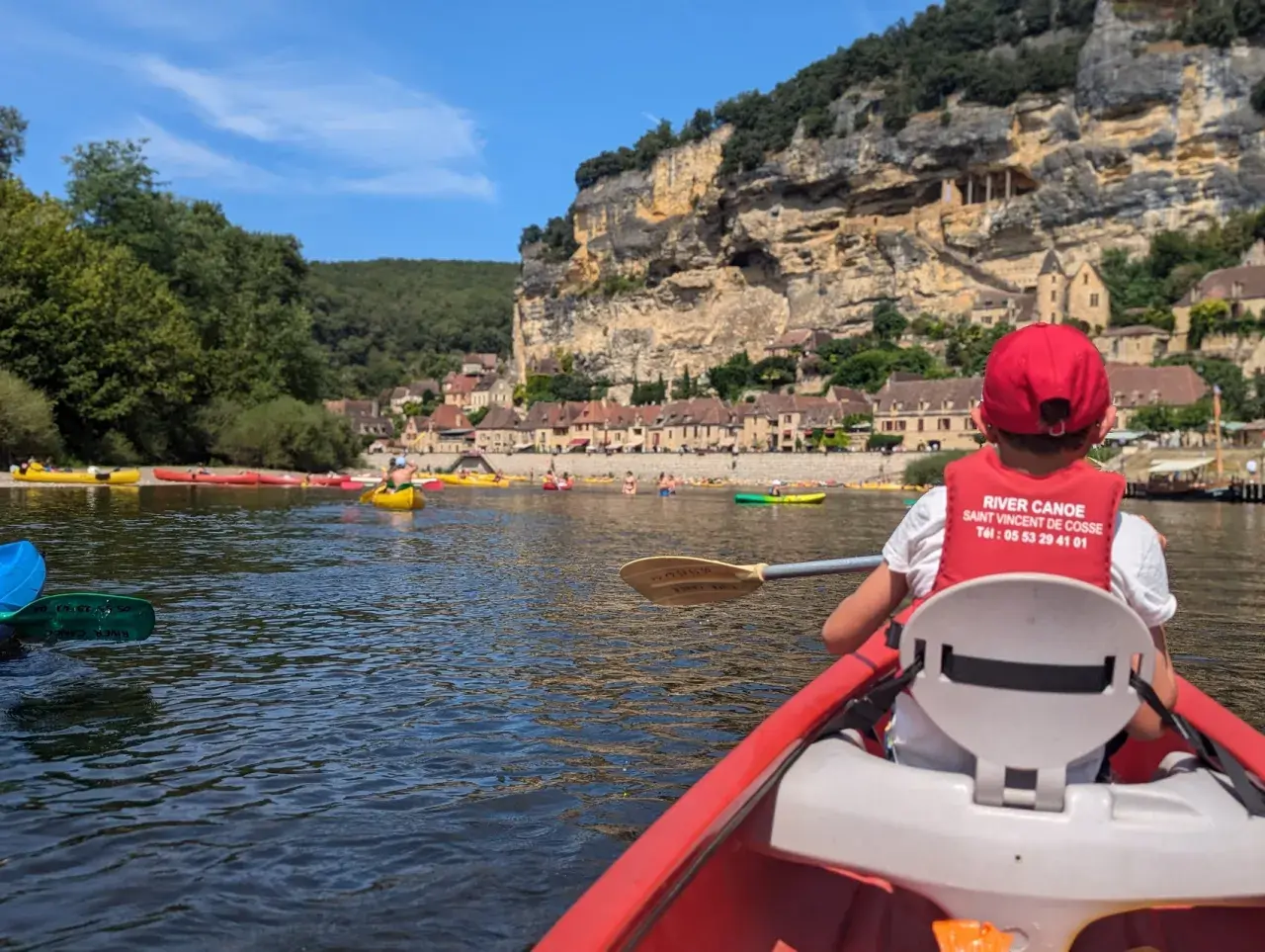 Enfant en canoë rouge, gilet de sauvetage "River Canoe", pour une visite insolite autour de Sarlat, avec des falaises et des maisons troglodytes en arrière-plan.