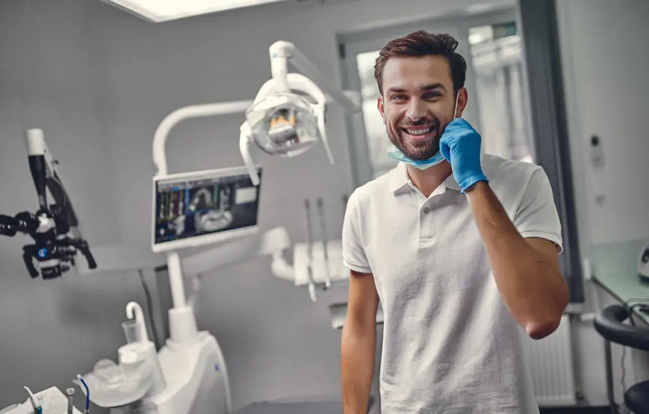 Uśmiechnięty dentysta w gabinecie, gotowy do pracy. Zastanawiasz się, ile trwa plombowanie zęba? To zależy od przypadku, ale zazwyczaj krótko!