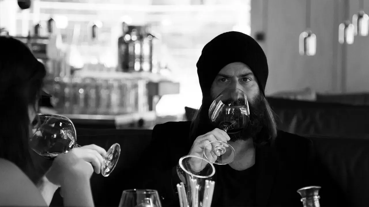 Ein Mann mit Bart und Mütze trinkt Wein. Er hält das Glas am Stiel, eine elegante Art, wie trinkt man wein.