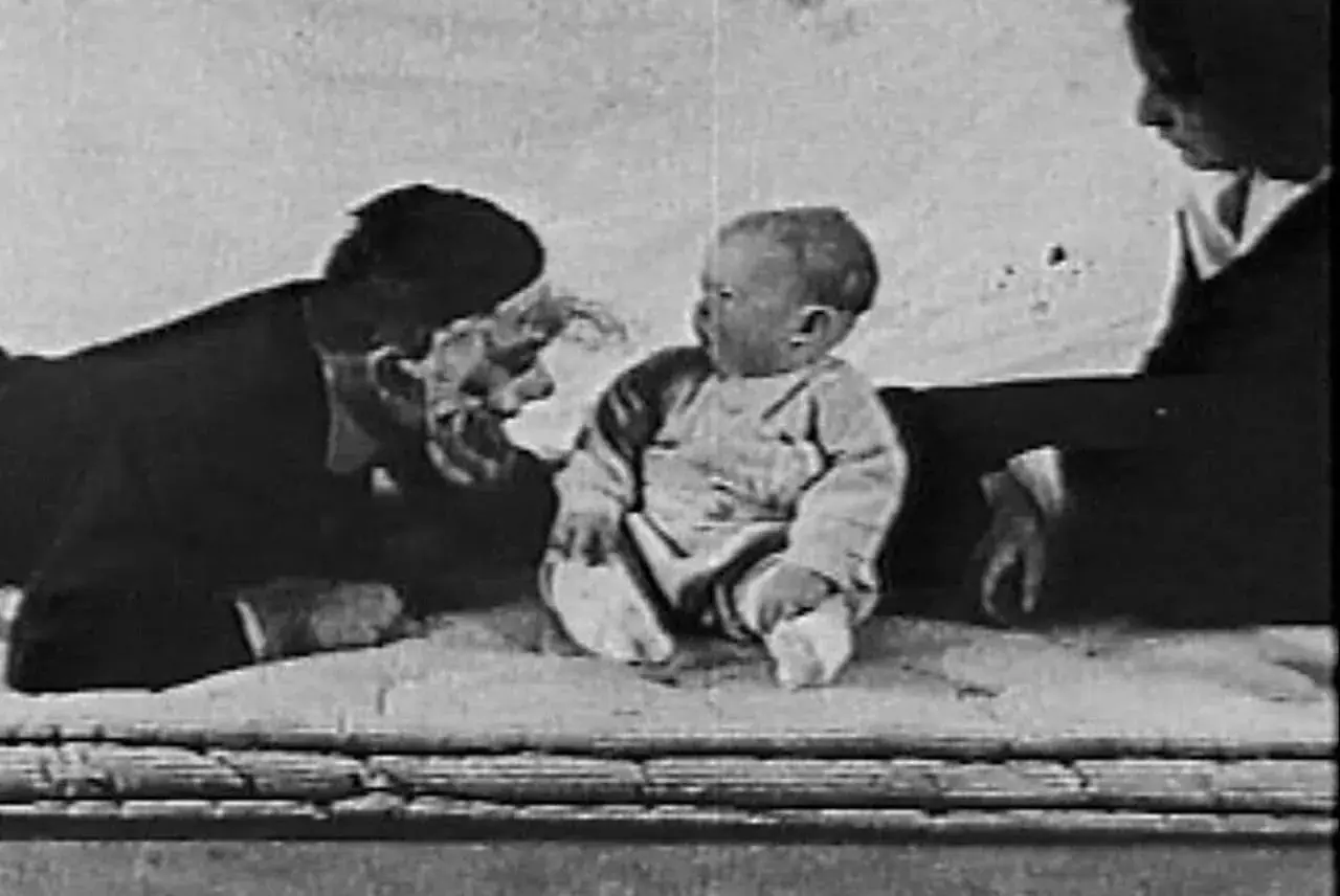 Bambino seduto, un uomo con maschera e una donna sullo sfondo, scena dall'esperimento piccolo Albert.