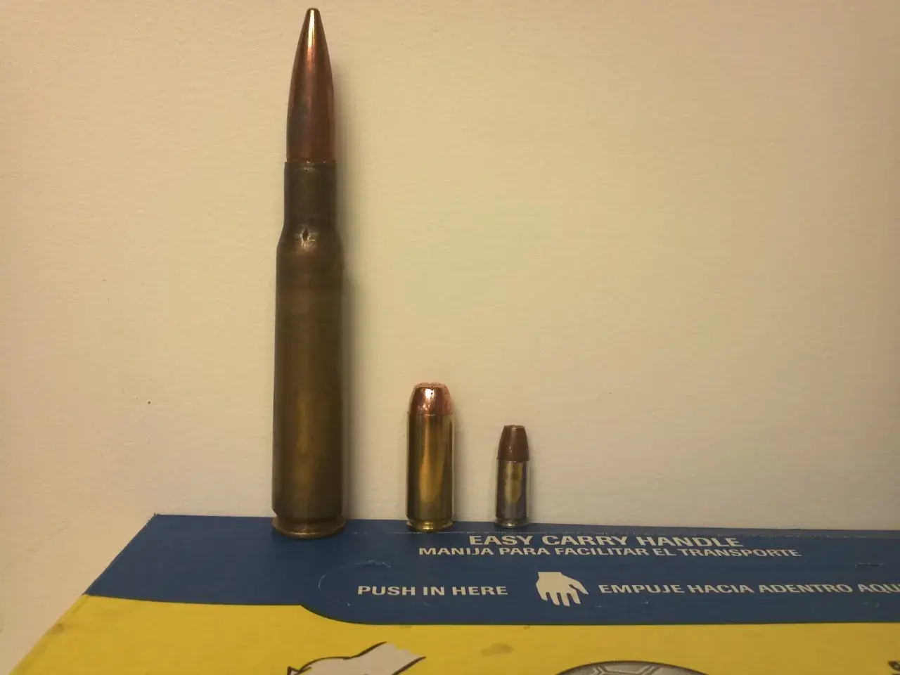 Trois cartouches de tailles différentes, dont une imposante de calibre 50 en mm, posées sur une boîte bleue et jaune.