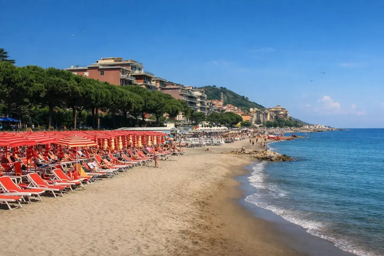 Plage d'Imperia animée, avec des rangées de parasols rouges et des chaises longues sur le sable. La mer bleue étincelle sous un ciel clair.